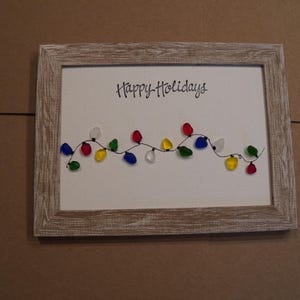 May include: A framed wall art piece featuring the text Happy Holidays in black script above a string of colorful sea glass pieces arranged to resemble Christmas lights in red, green, blue, yellow, and white, set in a light wood grain frame.