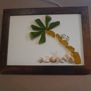 May include: A framed wall art piece featuring a palm tree constructed from green and amber sea glass shards on a white background. The base of the tree is surrounded by a collection of small, light-colored seashells and two white pebble-like accents.