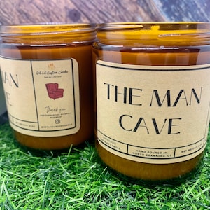 May include: Two amber glass candle jars with brown paper labels. The label on the left jar reads "The Man Cave" and the label on the right jar reads "The Man Cave". The labels have a rustic design and the text is in black.