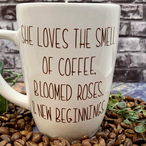 May include: White ceramic coffee mug with a brown inscription that reads "She loves the smell of coffee, bloomed roses, & new beginnings". The mug is sitting on a bed of coffee beans.