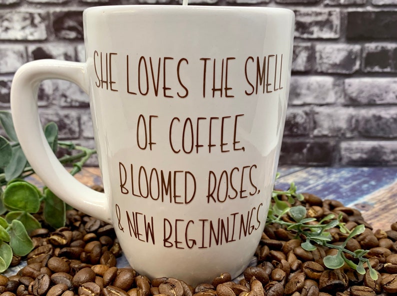 May include: White ceramic coffee mug with a brown inscription that reads "She loves the smell of coffee, bloomed roses, & new beginnings". The mug is sitting on a bed of coffee beans.