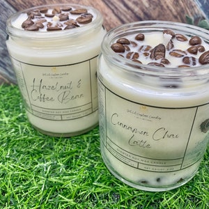 May include: Two glass jars filled with soy wax candles. The candles are topped with coffee beans. The jar on the left has a label that reads "Hazelnut & Coffee Bean" and the jar on the right has a label that reads "Cinnamon Chai Latte". Both jars are sitting on a green surface.