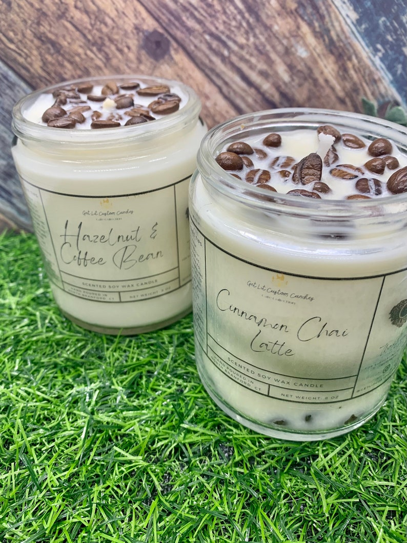 May include: Two glass jars filled with soy wax candles. The candles are topped with coffee beans. The jar on the left has a label that reads "Hazelnut & Coffee Bean" and the jar on the right has a label that reads "Cinnamon Chai Latte". Both jars are sitting on a green surface.