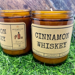 May include: Two amber glass candle jars with brown paper labels. The label on the left jar reads "MON KEY" and the label on the right jar reads "CINNAMON WHISKEY". The candle jars are sitting on a green grassy surface.