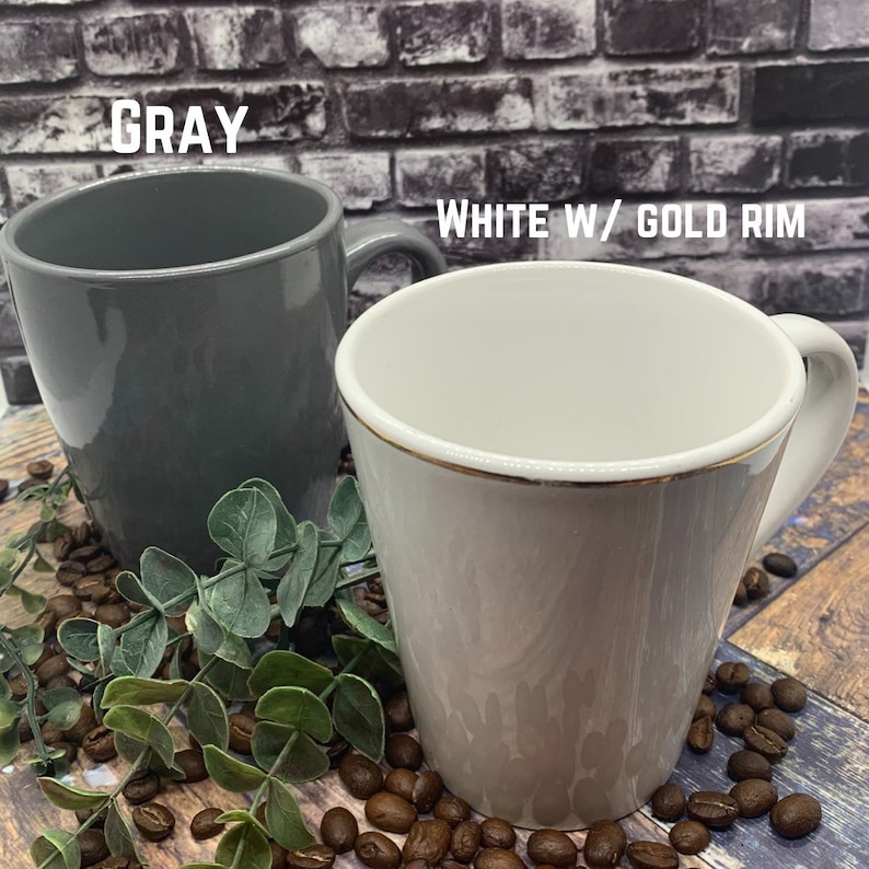 May include: Two coffee mugs, one gray and one white with a gold rim. The mugs are sitting on a wooden surface with coffee beans scattered around them.