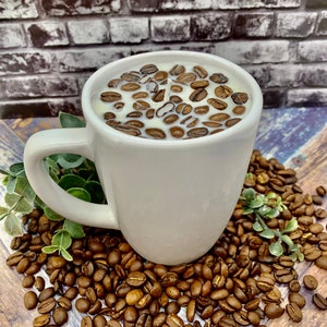 May include: White ceramic coffee mug candle with coffee beans embedded in the wax. The mug is sitting on a bed of coffee beans.