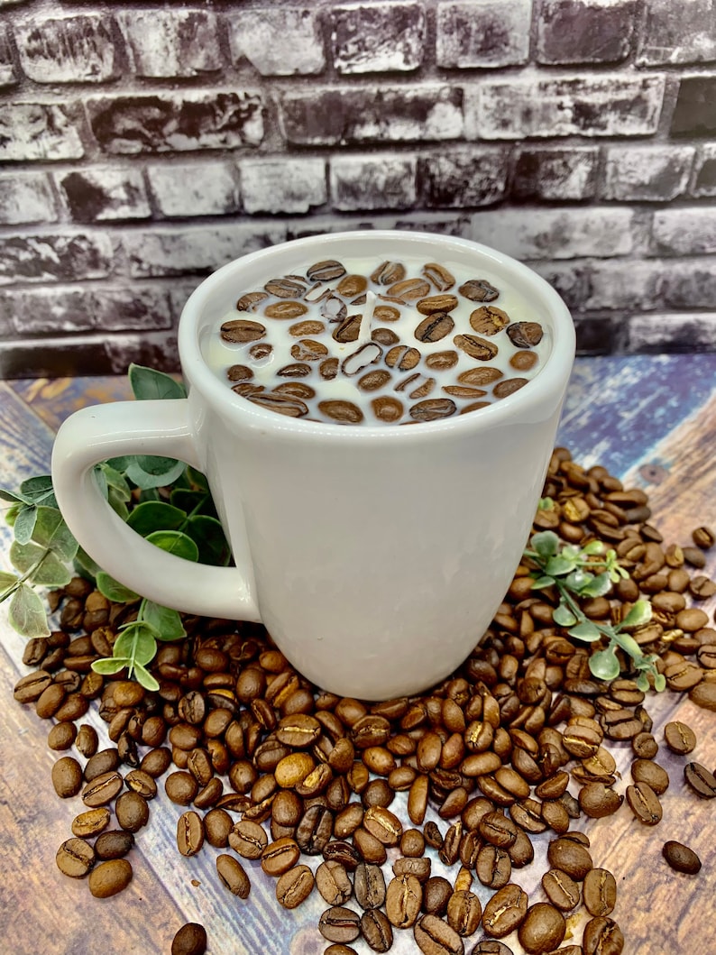 May include: White ceramic coffee mug candle with coffee beans embedded in the wax. The mug is sitting on a bed of coffee beans.
