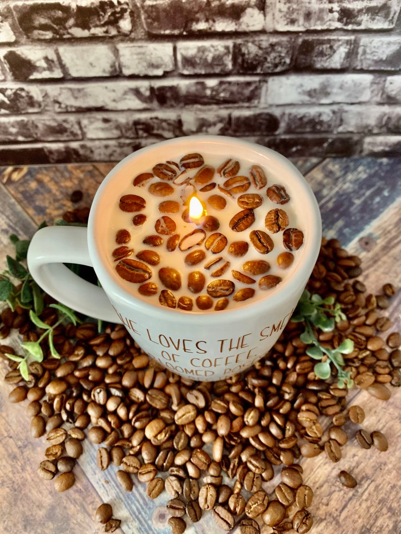 May include: A white ceramic coffee mug candle with a lit wick and coffee beans embedded in the wax. The mug has the text "He loves the smell of coffee" printed on the side. The mug is sitting on a bed of coffee beans.