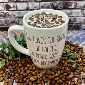 May include: White ceramic coffee mug candle with a brown coffee bean design. The mug has a handle and the text "She loves the smell of coffee, bloomed roses, & new beginnings" printed on the side.