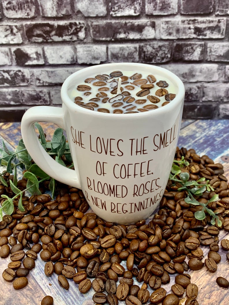 May include: White ceramic coffee mug candle with a brown coffee bean design. The mug has a handle and the text "She loves the smell of coffee, bloomed roses, & new beginnings" printed on the side.
