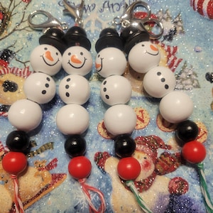 May include: Four snowman keychains made of white, black, and red wooden beads. Each keychain has a black top hat and a silver clip.