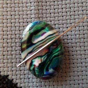 May include: An oval-shaped abalone shell bead with iridescent colors of green, blue, and purple, pierced by a long, thin, gold-colored needle. The bead rests on a gray cross-stitch fabric, suggesting a craft or jewelry-making theme.