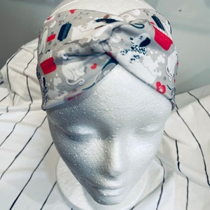 May include: A gray and white headband with a red and white pattern of dogs and bones. The headband is twisted and tied at the top.