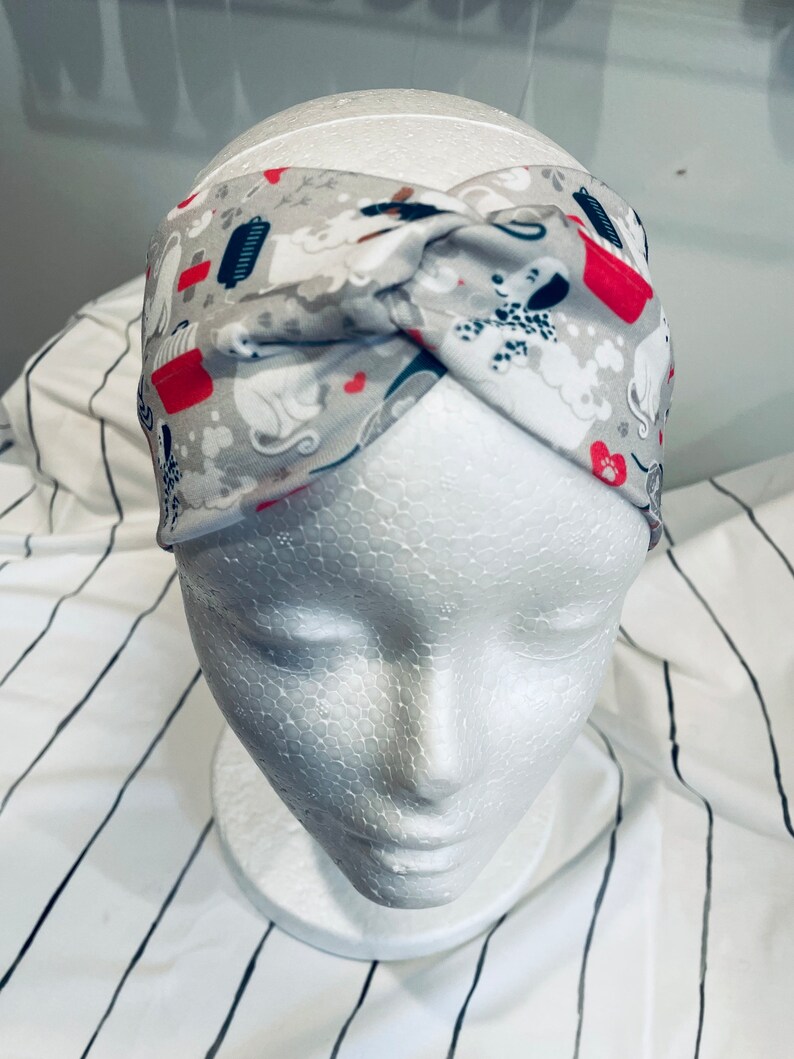 May include: A gray and white headband with a red and white pattern of dogs and bones. The headband is twisted and tied at the top.