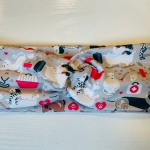 May include: A gray and white fabric headband with a pattern of dogs, cats, and pet-related items, including a red pet carrier, a bone, a paw print, and a first aid kit.