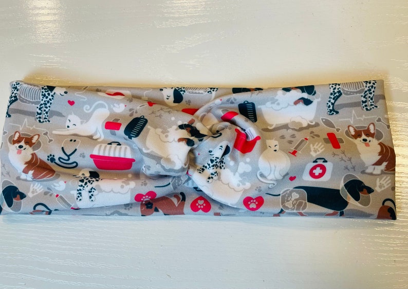 May include: A gray and white fabric headband with a pattern of dogs, cats, and pet-related items, including a red pet carrier, a bone, a paw print, and a first aid kit.