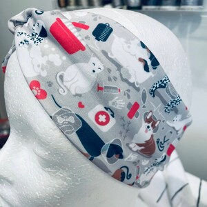 May include: A gray and white headband with a colorful pattern of dogs, cats, and other pet-related items. The pattern includes a red first aid cross, a red dog bone, and a red cat paw print.