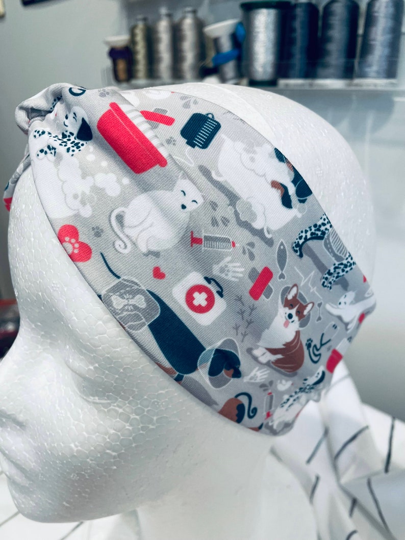 May include: A gray and white headband with a colorful pattern of dogs, cats, and other pet-related items. The pattern includes a red first aid cross, a red dog bone, and a red cat paw print.