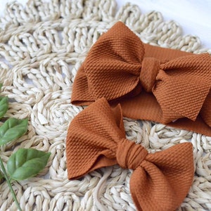 May include: Two brown fabric hair bows with a textured surface. The larger bow is on a headband. The bows are on a woven straw mat.