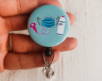 May include: A light blue round badge with a white face mask, a pink hand sanitizer bottle with the word "gel" on it, and a pink stethoscope. The badge is attached to a black retractable badge reel with a clear plastic clip.