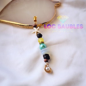 May include: A gold charm with a white seashell and a pearl hanging from a string of beads. The beads are yellow, green, black, and brown. The charm is labeled "LOC BAUBLES handcrafted accessories".