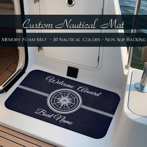 May include: A navy blue memory foam mat with a nautical theme. The mat features a compass rose design and the words "Welcome Aboard" and "Boat Name" in white script. The mat is designed for boats.
