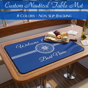 May include: A rectangular blue nautical table mat featuring a white compass rose graphic, rope border details, and the text "Welcome" and "Boat Name". The mat is displayed on a wooden table with a wooden serving board of cheese and grapes.