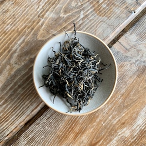 May include: A small white bowl filled with loose leaf black tea with golden tips.