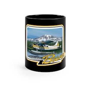 Puede incluir: Taza de café negra con un gráfico de un hidroavión De Havilland Beaver amarillo y blanco en un lago frente a una montaña. El texto "De Havilland" y "Beaver" están en amarillo en la taza.