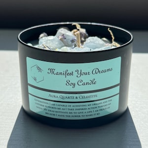 May include: A black soy candle in a metal container, filled with clear quartz and other crystals. The label reads "Manifest Your Dreams Soy Candle" with the text "Aura Quartz & Celestite" and an affirmation.