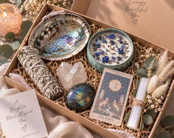 Puede incluir: Una caja de regalo que contiene una concha de abulón iridiscente, una vela con cristales azules y flores secas, un atado de salvia, un grupo de cuarzo transparente, una piedra de labradorita oscura, una carta del tarot y una varita de selenita.