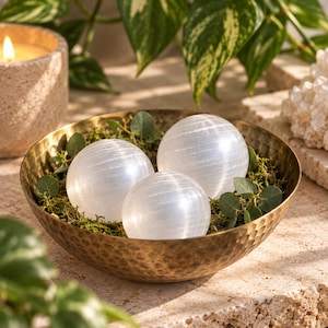 May include: Three smooth, white, spherical selenite crystals are nestled inside a round, hammered brass bowl. The bowl contains green moss and small green leaves tucked around the base of the three polished stone spheres, creating a display.