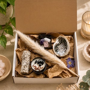 Puede incluir: Una caja de regalo de cartón que contiene un atado de salvia, una vela negra pequeña con piedras negras y pétalos secos, una concha de abulón pulida, tres cristales pequeños en bruto, un penacho de hierba de la pampa y una flor morada.