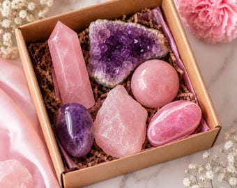 May include: A square cardboard gift box filled with brown paper crinkle shred containing six assorted crystals: a tall rose quartz point, a purple amethyst cluster, a smooth rose quartz sphere, a polished purple amethyst egg, a raw rose quartz chunk, and a smooth pink rose quartz stone.