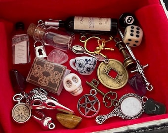 May include: A rectangular jewelry box with a red velvet interior containing a collection of miniature charms and trinkets, including a wine bottle, dice, a skull, a pocket watch, a mirror, a pentagram, keys, a spoon, and various crystals.