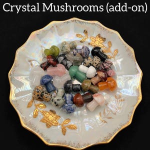 May include: A collection of crystal mushroom figurines in various colors, including green, pink, and black, are displayed in a decorative, gold-rimmed dish. The dish has a white and gold floral design. The text "Crystal Mushrooms (add-on)" is at the top.