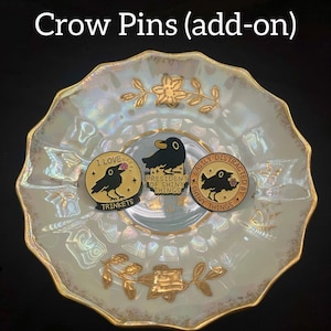 May include: Three enamel pins featuring crow illustrations. One pin reads "I love trinkets", another says "President of shiny things", and the third says "Easily distracted by shiny things". The pins are gold, black, and pink. The pins are displayed on a decorative plate.