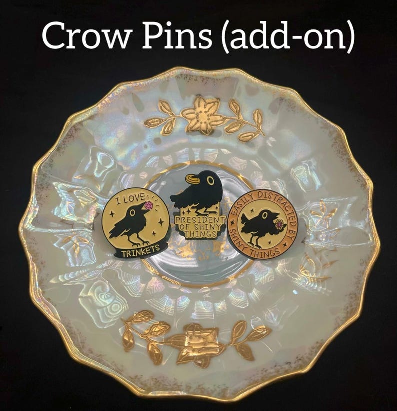 May include: Three enamel pins featuring crow illustrations. One pin reads "I love trinkets", another says "President of shiny things", and the third says "Easily distracted by shiny things". The pins are gold, black, and pink. The pins are displayed on a decorative plate.