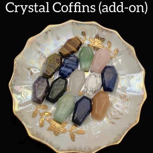 May include: A collection of crystal coffin-shaped stones in various colors, including green, pink, blue, and brown. The crystals are arranged on a decorative, scalloped-edge dish with gold accents. Text at the top reads "Crystal Coffins (add-on)".