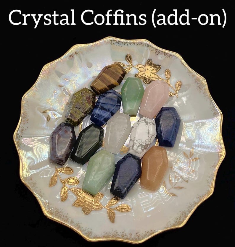 May include: A collection of crystal coffin-shaped stones in various colors, including green, pink, blue, and brown. The crystals are arranged on a decorative, scalloped-edge dish with gold accents. Text at the top reads "Crystal Coffins (add-on)".