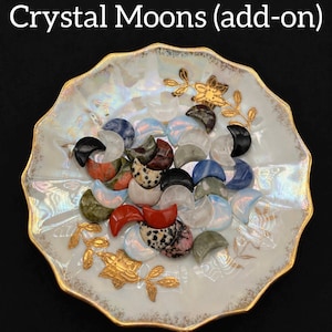 May include: A collection of crystal moon-shaped stones in various colors, including black, red, blue, and white, arranged on a decorative, scalloped-edge plate with gold accents. The plate has a pearlescent finish and gold floral designs. The text "Crystal Moons (add-on)" is at the top.