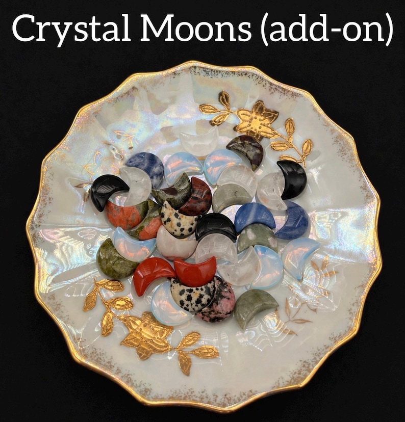 May include: A collection of crystal moon-shaped stones in various colors, including black, red, blue, and white, arranged on a decorative, scalloped-edge plate with gold accents. The plate has a pearlescent finish and gold floral designs. The text "Crystal Moons (add-on)" is at the top.
