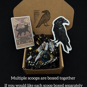 May include: A cardboard box containing a collection of items, including a tarot card with a black cat, stickers of ravens, and various small objects. The box is lined with black fabric with gold stars. Text on the image reads: "Multiple scoops are boxed together."