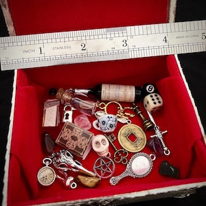 May include: A red velvet-lined box filled with miniature items. Includes a small wine bottle, dice, a skull, a book, a key, a mirror, and other charms. A stainless steel ruler measures the box, showing it is approximately 4 inches wide.