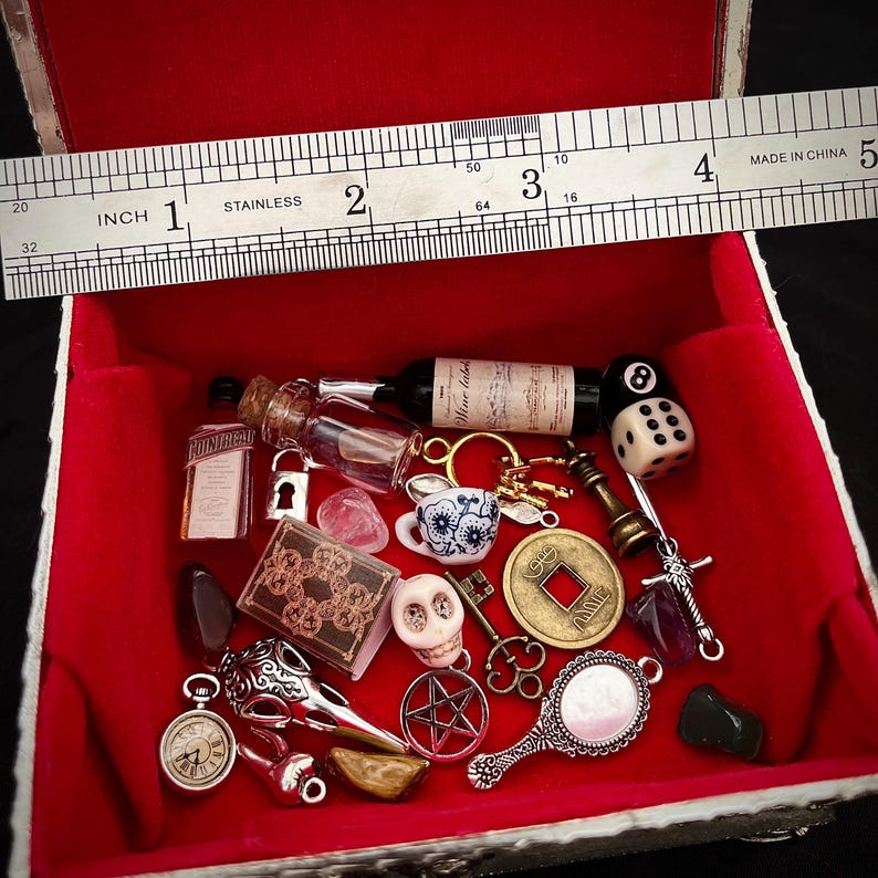 May include: A red velvet-lined box filled with miniature items. Includes a small wine bottle, dice, a skull, a book, a key, a mirror, and other charms. A stainless steel ruler measures the box, showing it is approximately 4 inches wide.