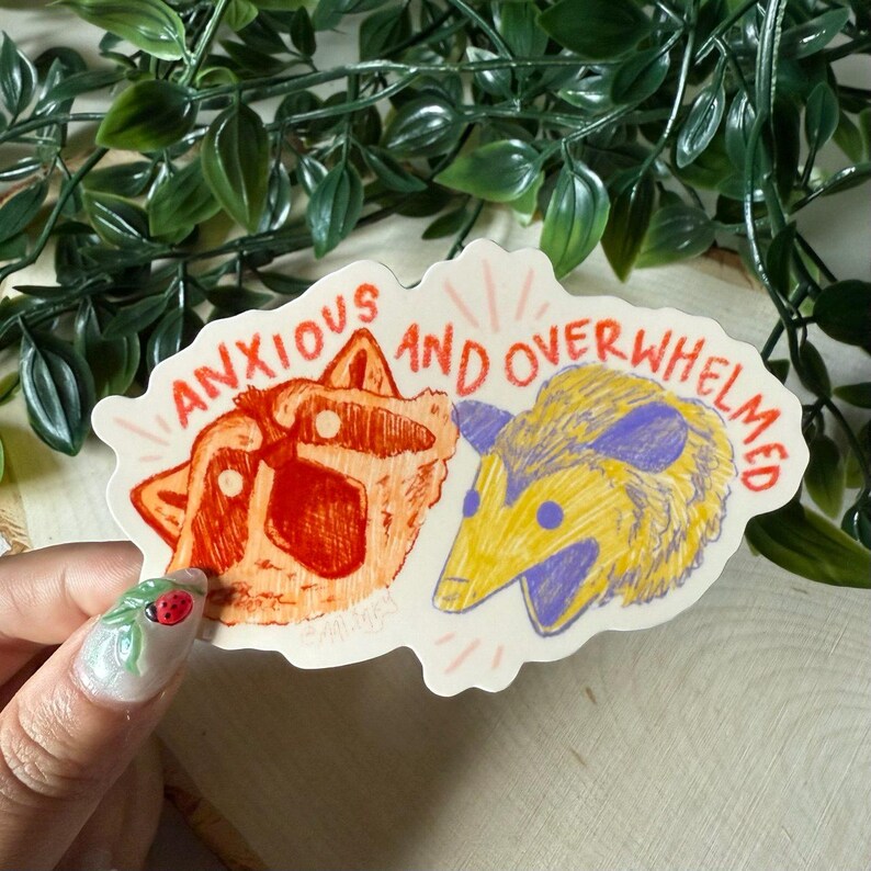 May include: A sticker with a raccoon and an opossum with the text "anxious and overwhelmed". The raccoon is red and the opossum is yellow.