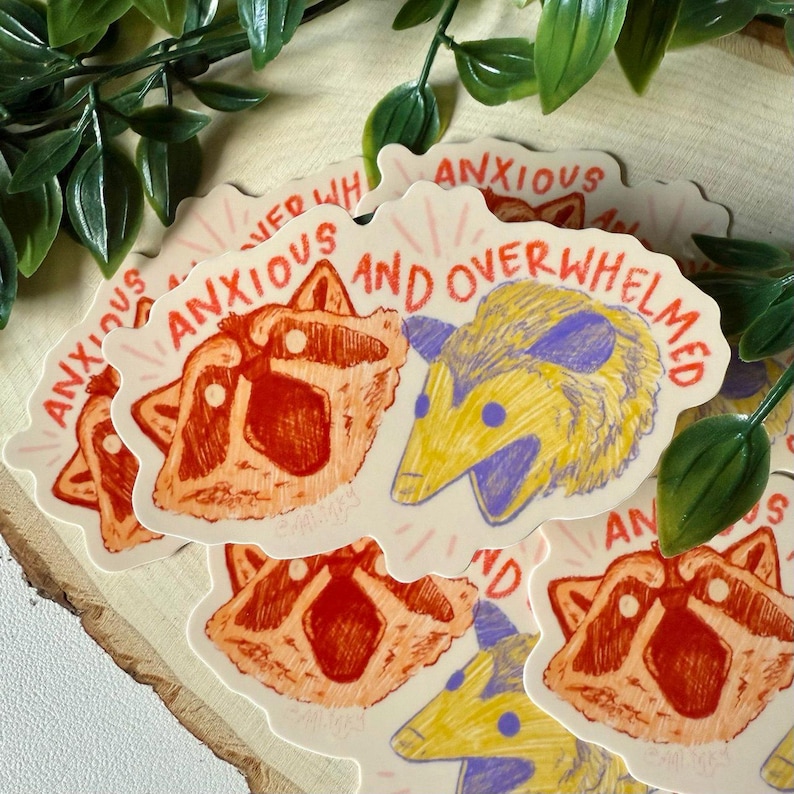 May include: A set of four stickers featuring a raccoon and a possum with the text "Anxious and Overwhelmed". The stickers are on a wooden surface with green leaves.