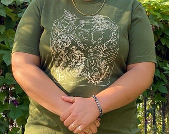 May include: A green t-shirt with a white line drawing of a skull surrounded by flowers and plants. The text "Model wearing XL" is at the bottom of the image.
