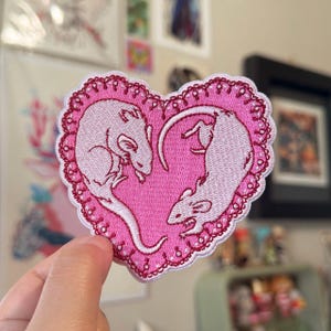 May include: A heart-shaped embroidered patch featuring two white rats on a pink background. The patch has a decorative, scalloped edge with red stitching. The rats are positioned to form a heart shape.