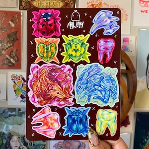 Puede incluir: Una hoja de pegatinas con una variedad de diseños coloridos, incluyendo una mariquita, un diente, un escarabajo y un conejo. Las pegatinas están sobre un fondo rojo oscuro y tienen un borde blanco. El texto "MAL.inky" está impreso en blanco en la hoja.